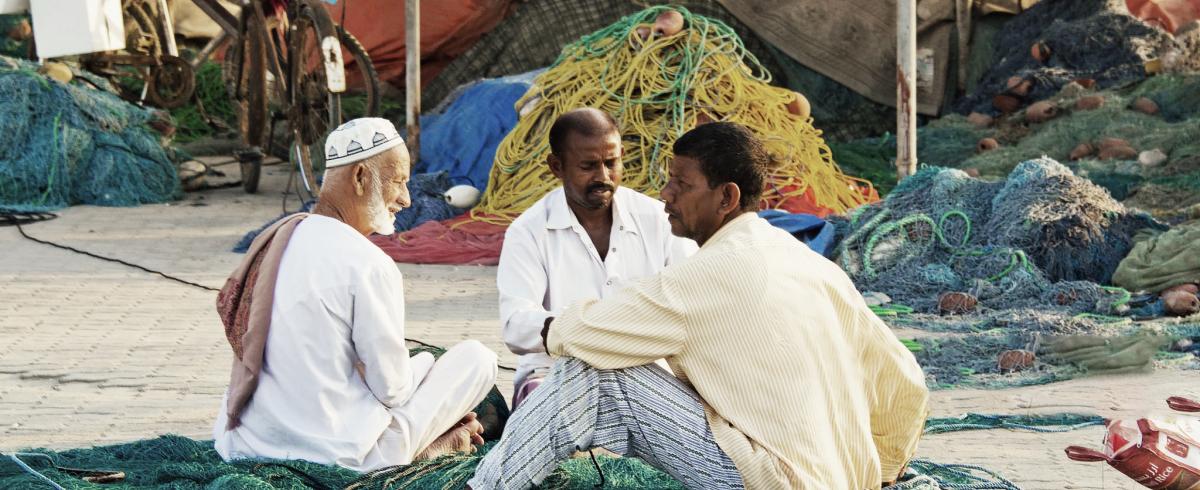 3 fishermen sitting on a beach in front of their fishing boats.